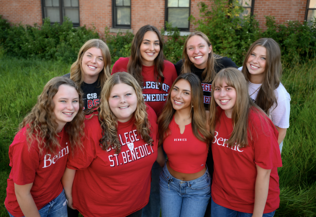 A group of nine young women stand together outside, smiling at the camera. They are wearing red and blue college shirts, with greenery and a brick building in the background.